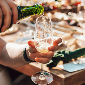 Person pouring green wine into a glass on a wooden table with a blurred outdoor background