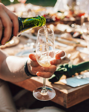Person pouring green wine into a glass on a wooden table with a blurred outdoor background