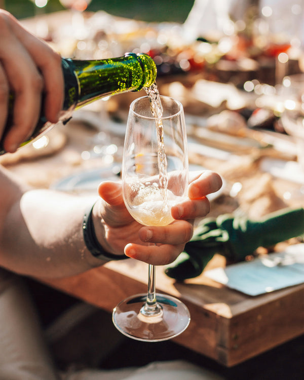 Person pouring green wine into a glass on a wooden table with a blurred outdoor background